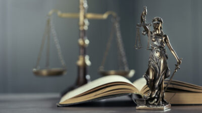 Statue einer Justitia. Im Hintergrund ein aufgeklapptes Buch und eine rechtschaffende Waage.