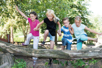 Vier Kinder springen Hand in Hand über einen Baumstamm.