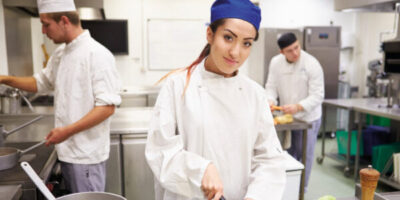Junge Frau steht in einer Großküche und schneidet Karotten. Im Hintergrund sind zwei männliche Küchenhilfen zu erkennen.