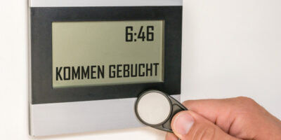 Digitales Anzeigeschild zeigt Uhrzeit und liest KOMMEN GEBUCHT. Eine Hand hält einen Transponder vor das Anzeigeschild.